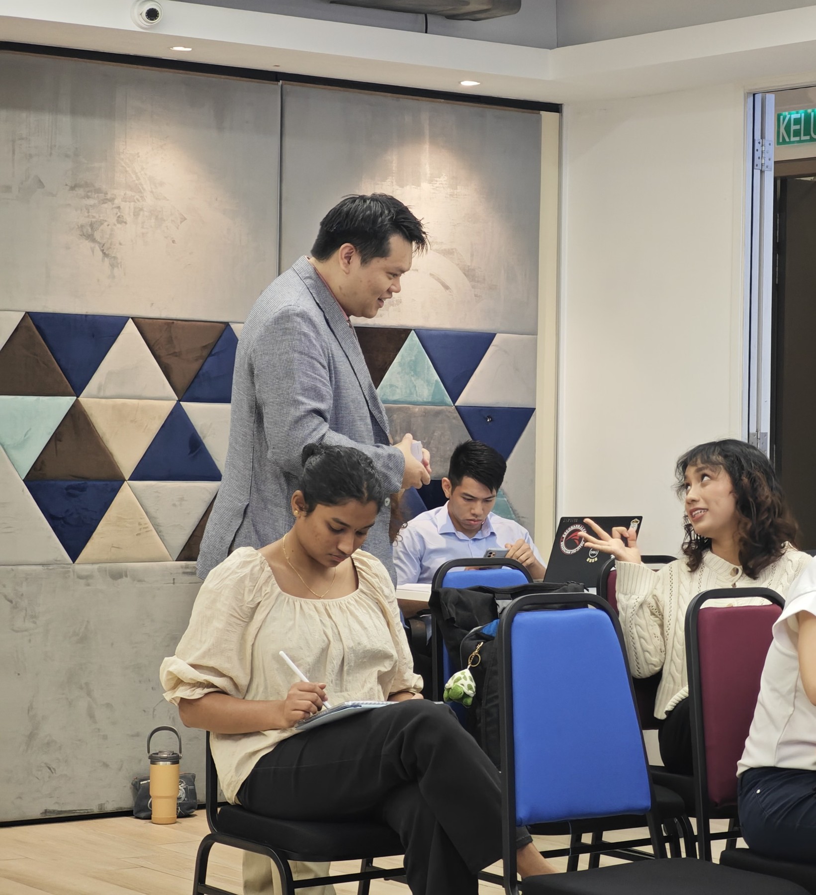 Aaron standing among seated participants, smiling as one of them asks a question while another writes notes on a tablet