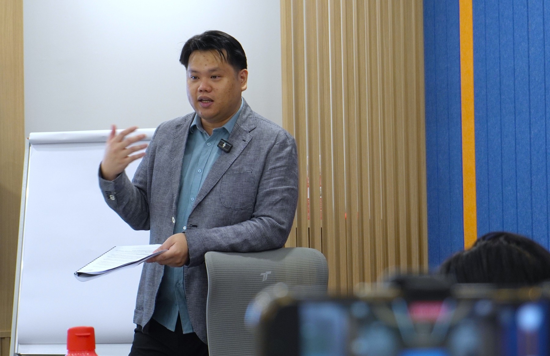 Aaron Cheah holding printed notes and gesturing as he speaks beside a flip chart, with participants in the foreground